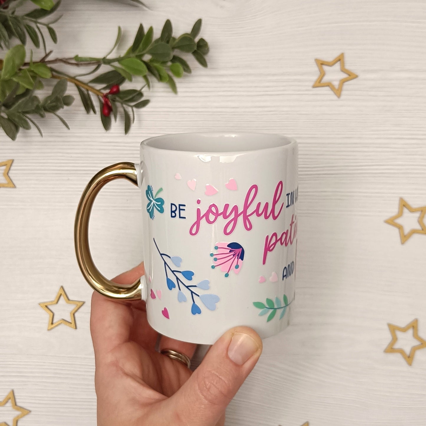 White mug with 'Be joyful in hope, patient in affliction and faithful in prayer' text held by a hand against a decorative background with stars and greenery.
