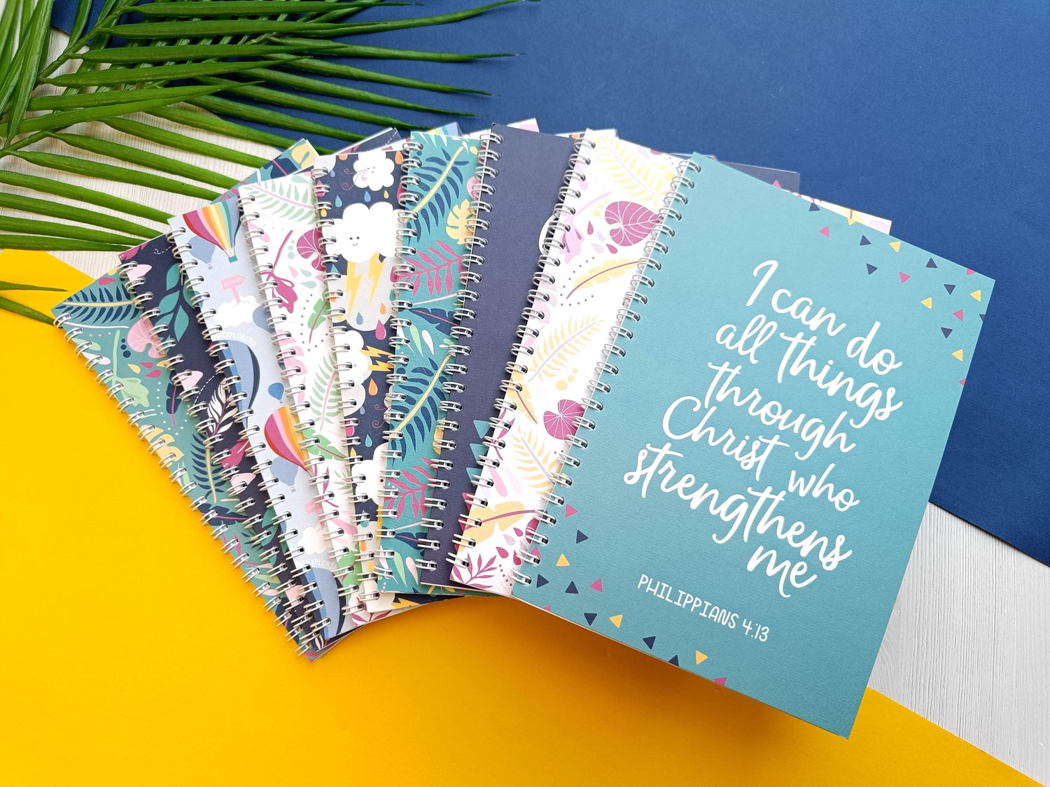 9 spiral bound notebooks, fanned out on top of each other. Bright, colourful covers with scripture verses on some of them.