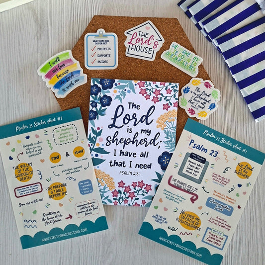 A styled flat-lay of Bible-themed sticker sheets and a central floral card reading 'The Lord is my shepherd I have all that I need PSALM 23:1' on a light wood surface, with a cork board, blue-and-white striped fabric, and other stationery props around it.